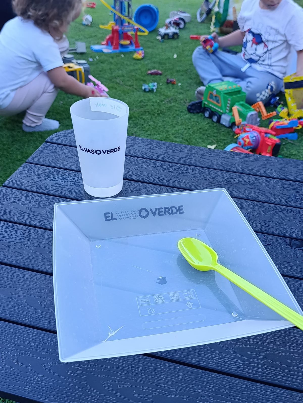 Vaso y plato reutilizables de El Vaso Verde sobre una mesa, con niños jugando al fondo durante un cumpleaños en el parque.